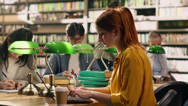 Red-haired woman student drinking coffee while using laptop at library desk, combining academic focus of writing assignment with small study break.