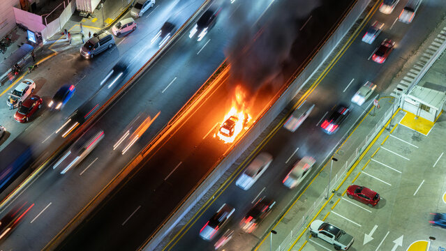 Aerial view of a car engulfed in flames on a highway at night with traffic moving in surrounding lanes, Dominican Republic.