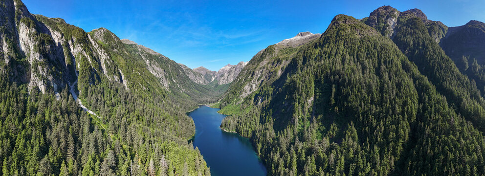 Aerial view of a deep blue mountain lake nestled within a dense coniferous forest and steep rocky slopes under a clear blue sky in Sitka, Alaska, United States.