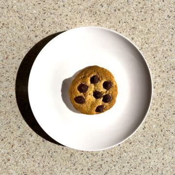 High contrast overhead view of a chocolate chip cookie on a white plate sitting on a benchtop