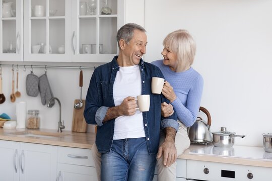 Smiling Elderly Caucasian Man And Woman Holding Coffee Cups While Sitting Together In Bright Modern Kitchen, Enjoying Each Other's Company. Warm Family Bonding Moment At Home.