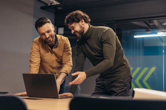 Happy Caucasian Men Engaged In Collaborative Work While Smiling At Laptop In Modern Office Setting. They Are Sharing Ideas And Working Together At A Meeting Table, Business Concept.