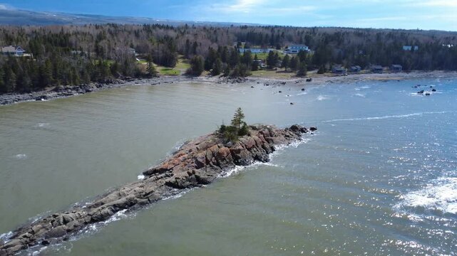 Drone footage of a rocky coastal islet topped with evergreen trees, surrounded by calm waters and a forested shoreline with scattered homes in Quebec, Canada.