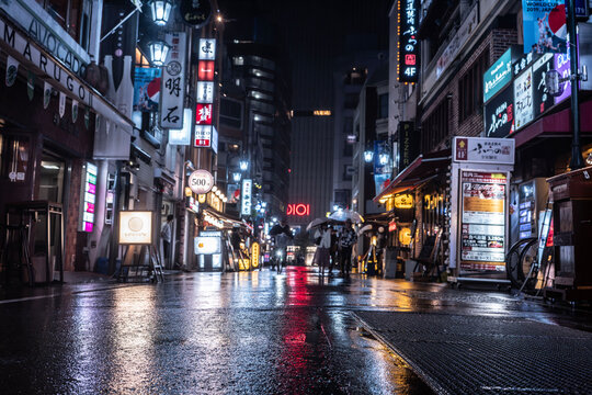 雨の新宿三丁目・飲食店街のネオンとマルイの赤いロゴが路面に映える夜景