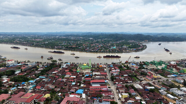 Aerial view of the Berau River with coal barges and tugboats navigating past a green-roofed mosque and dense residential neighborhoods in Berau Regency, East Kalimantan, Indonesia.