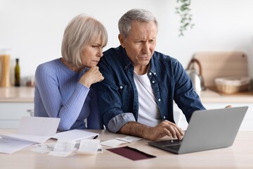 Worried Elderly Caucasian Couple Sitting At Kitchen Table, Reviewing Bills And Financial Documents...