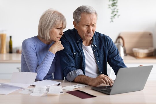 Worried Elderly Caucasian Couple Sitting At Kitchen Table, Reviewing Bills And Financial Documents Together, Expressing Concern Over Household Finances. Concept Of Financial Strain And Planning.