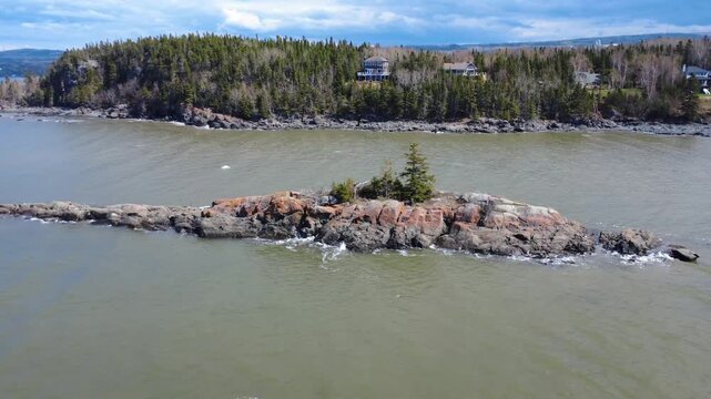 Drone footage descending toward a rocky islet with evergreen trees, surrounded by calm bay waters and a forested shoreline near Metis-sur-Mer, Quebec, Canada.