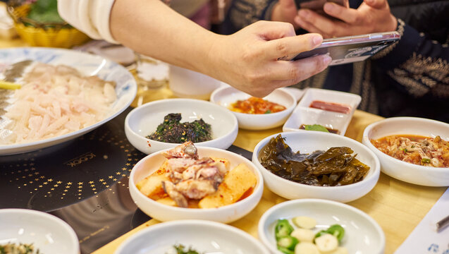 These images capture a diner using a smartphone to photograph a rich spread of Korean coastal delicacies, including sashimi and traditional banchan. It beautifully highlights modern digital dining cul