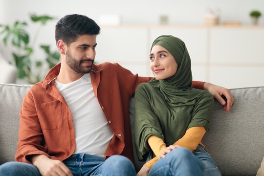 Happy Young Arab Couple Sitting Together On A Sofa, Smiling At Each Other In Their Cozy Home. Enjoying Quality Time In A Relaxed Setting Together, Indicating Love And Affection, Lifestyle Concept.
