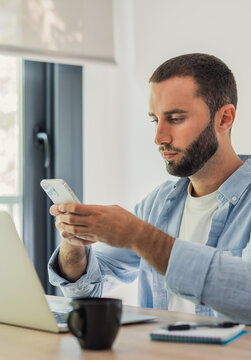 Young man focused on connecting and communicating with smartphone, sitting at desk with laptop, embodying modern remote work and digital lifestyle