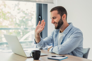 Obraz premium Man smiling and waving to a laptop screen while participating in an online video call from his bright home office, connecting remotely