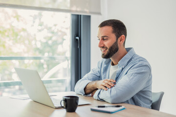 Naklejka premium Young man smiling at his laptop while telecommuting, working remotely from a modern home office with a cup of coffee and a notebook on a wooden desk 