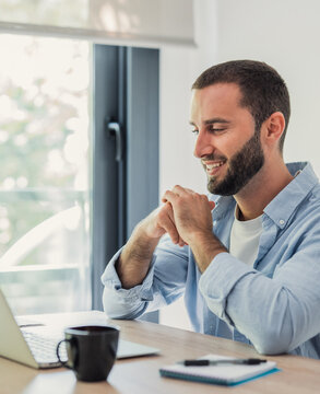 Bearded man smiling and looking at a laptop screen on a wooden desk, enjoying flexible work from home while connecting online, with a coffee mug and notebook nearby