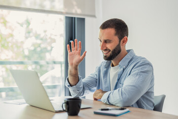 Obraz premium Man smiling and waving hand at a laptop screen, engaging in a virtual video call at home, symbolizing remote work and modern communication