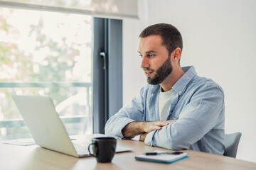 Naklejka premium Young man concentrating on laptop screen while sitting at desk in a modern home office space, working or studying remotely with natural light from a window