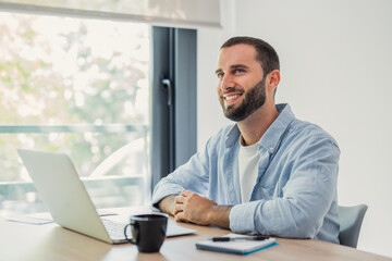 Fototapeta premium Young man telecommuting from a well-lit home office, smiling and looking out of the window while working on a laptop, representing modern remote work