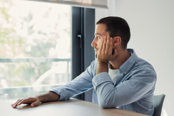 Obraz premium Young man sitting at a table at home. Thoughtfully looking out the window. Embracing introspection as he considers his future plans or past events in a moment of quiet reflection and mental processing