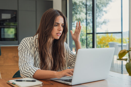 Young woman working on her laptop from home, experiencing frustration and confusion due to a technical problem or difficult task, raising her hand in exasperation and stress