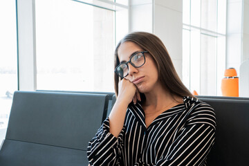 Fototapeta premium Woman in glasses and striped shirt resting head on hand, showing expressions of sadness, tiredness, and boredom while waiting for a flight at an airport terminal