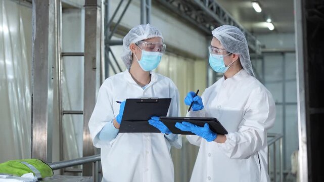 Quality control team using digital tablet and clipboard for production line audit in modern plant, Female engineers wearing face masks and safety goggles working in high-standard processing facility