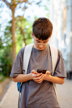 Vertical shot of teen student with backpack uses smartphone outdoors on sunny city street. Concept of mobile communication, online learning, school life, social media and digital lifestyle.