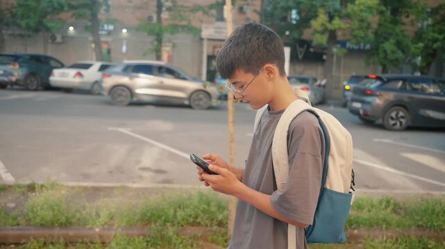 Teen student with backpack checks smartphone near city road. Concept of navigation app, location sharing, school commute, urban safety and independent travel.