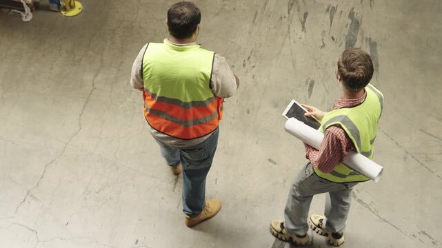 Construction professionals collaborate on blueprints and logistics planning within dark warehouse setting, utilizing tablets and detailed construction drawings during project inspection.