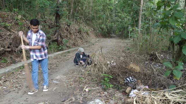Near-start to middle frame shows a man in a plaid shirt sweeping a dirt path while a woman follows with a bag, set on a sunlit forest trail