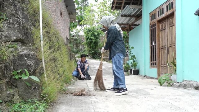 The full clip follows a daytime outdoor cleaning sequence: a woman sweeps a concrete path beside a turquoise house, with a man visible in the background
