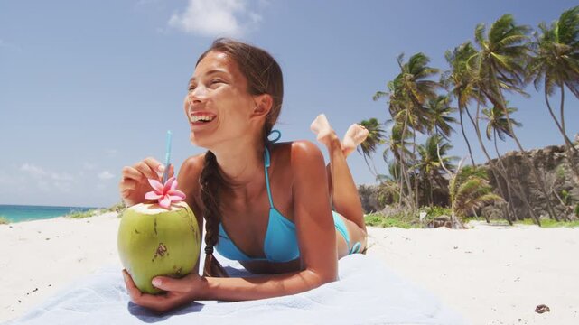 Happy beach bikini woman relaxing drinking fresh coconut water lying down sunbathing on fun Caribbean vacation tanning under the tropical sun. Video is unretouched and model without makeup.