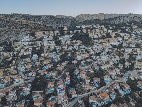 Aerial view of residential houses with swimming pools nestled on a hillside with winding roads and a rocky mountain backdrop in Calp, Valencian Community, Spain.