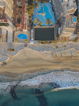 Aerial view of a coastal resort with swimming pools, a patterned promenade, and sandy beach meeting the turquoise sea in Calp, Valencian Community, Spain.
