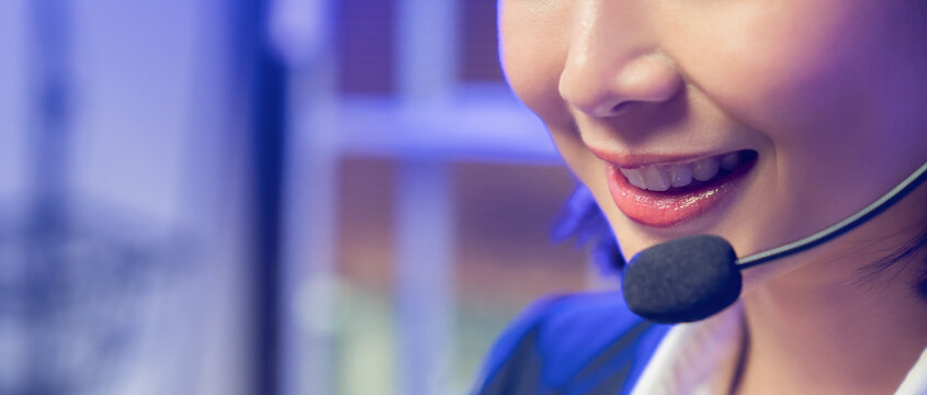 Smiling young Asian customer service representative wearing microphone headset and working on laptop in modern office.