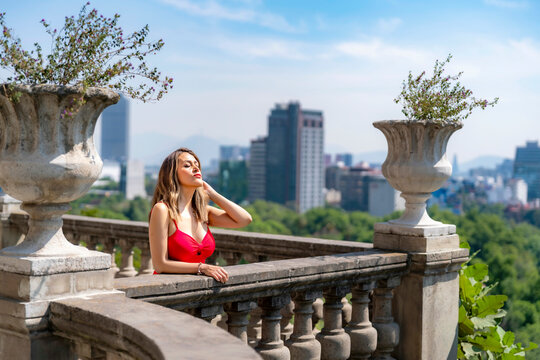 Young smiling latina woman posing calmly and thoughtfully on a balcony while enjoying the view toward Reforma avenue at Castillo de Chapultepec in Mexico City, wearing a vibrant elegant red dress