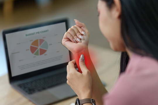 Asian woman holding painful wrist with red glow from carpal tunnel syndrome at laptop