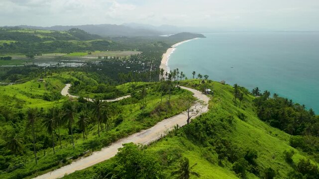 Aerial forwards along winding hilltop road leading toward Bato ni Ningning in San Vicente Palawan with lush green hills, coastal town and sandy beach