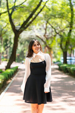 Young smiling latina woman posing relaxed, cheerful outdoors at Parque Mexico in Condesa, Ciudad de Mexico enjoying a sunny day surrounded trees and soft light, wearing a black dress with white blouse