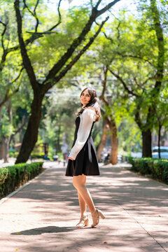 Young smiling latina woman twirling playfully, cheerful and carefree outdoors at Parque Mexico in Condesa, Ciudad de Mexico surrounded by trees sunlight, wearing a black dress, white blouse in motion