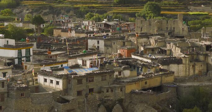 Kagbeni, Mustang Nepal, lateral tracking shot focusing on traditional mud brick architecture and damaged historic stone houses nestled beneath hills, surrounded by agricultural land.