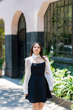 Young smiling latina woman posing relaxed, cheerful on streets near Parque Mexico in Condesa, Ciudad de Mexico surrounded by white architecture greenery, wearing a black dress, white blouse, sunlight