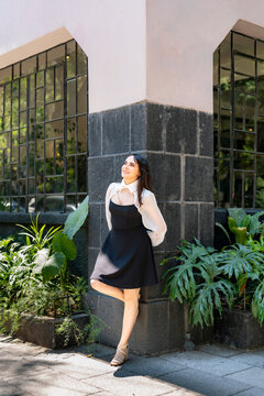 Young smiling latina woman posing relaxed, cheerful on streets near Parque Mexico in Condesa, Ciudad de Mexico surrounded by white architecture greenery, wearing a black dress, white blouse, sunlight