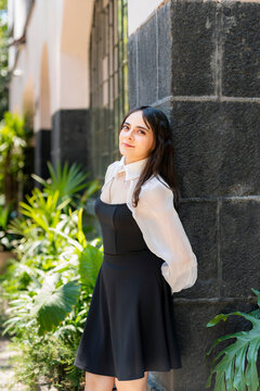 Young smiling latina woman posing relaxed, cheerful on streets near Parque Mexico in Condesa, Ciudad de Mexico surrounded by white architecture greenery, wearing a black dress, white blouse, sunlight