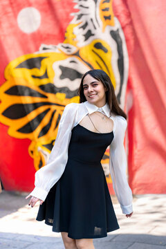 Young smiling latina woman posing relaxed, cheerful on streets near Parque Mexico in Condesa, Ciudad de Mexico surrounded by white architecture greenery, wearing a black dress, white blouse, sunlight