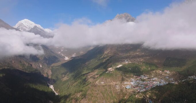 Slow gliding drone shot of Namche Bazaar, Nepal, with a low cloud layer cuts across the frame, separating the deep valley from the towering sunlit peaks of Thamserku and Ama Dablam above.
