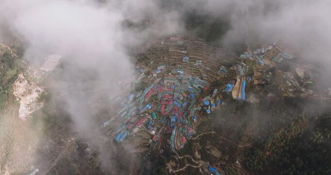 Namche Bazaar, Khumbu Nepal, spectacular high-angle aerial shot capturing the iconic horseshoe-shaped mountain village settlement, green hillside, and muddy slopes through drifting clouds