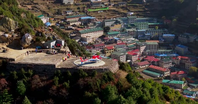 Namche Bazaar, Everest, Nepal. Orbiting drone shot looking down at a landed helicopter on the hilltop helipad as passengers step out, revealing the colorful hillside lodges and green hills