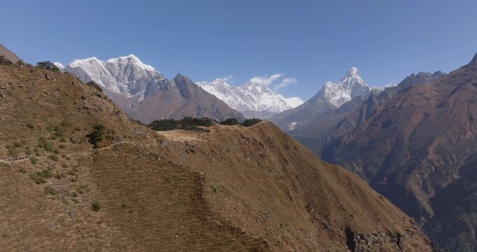 Drone glide in toward a panoramic view of Taboche, Mount Everest, Lhotse, and Ama Dablam rising above mountain trails, also showing rugged muddy terrain and green hills