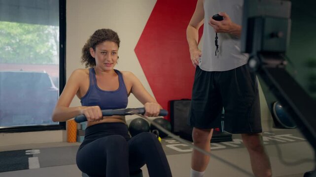 Young athletic woman using rowing machine with trainer in fitness gym. 