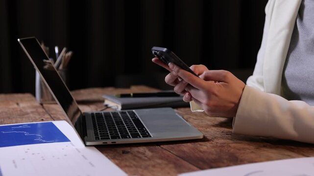 Businesswoman multitasking using smartphone and laptop at desk. Business professional sitting at a wooden desk, synchronizing data between her laptop and smartphone while working on financial charts.